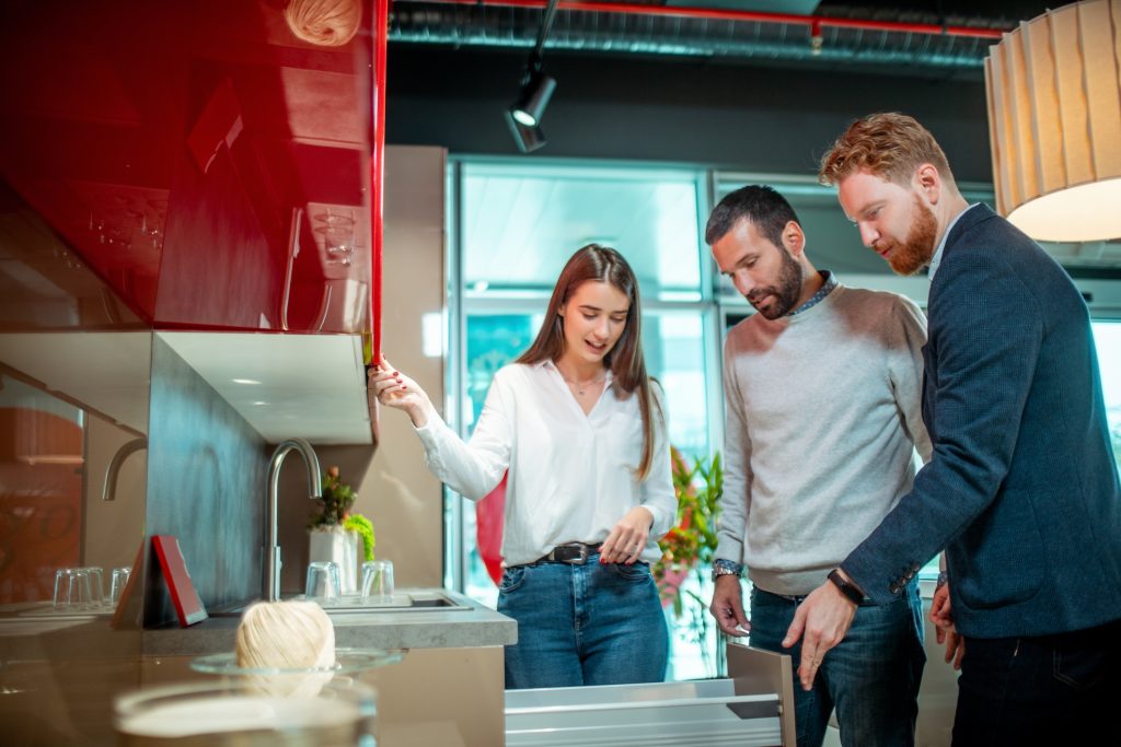 A store clerk demonstrating some kitchen design ideas and opening a cabinet drawer for a young couple in a kitchen equipment store